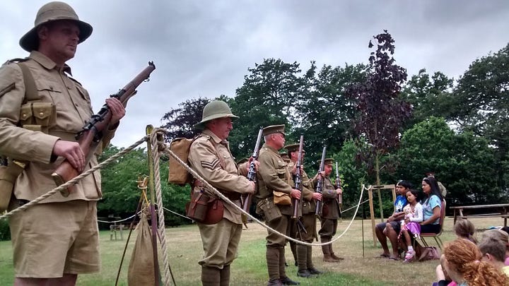 Man in WWI uniform holding open tin to reveal pack of tobacco and pack of cigarettes, men lined up with rifles in WWI uniform with modern kids watching