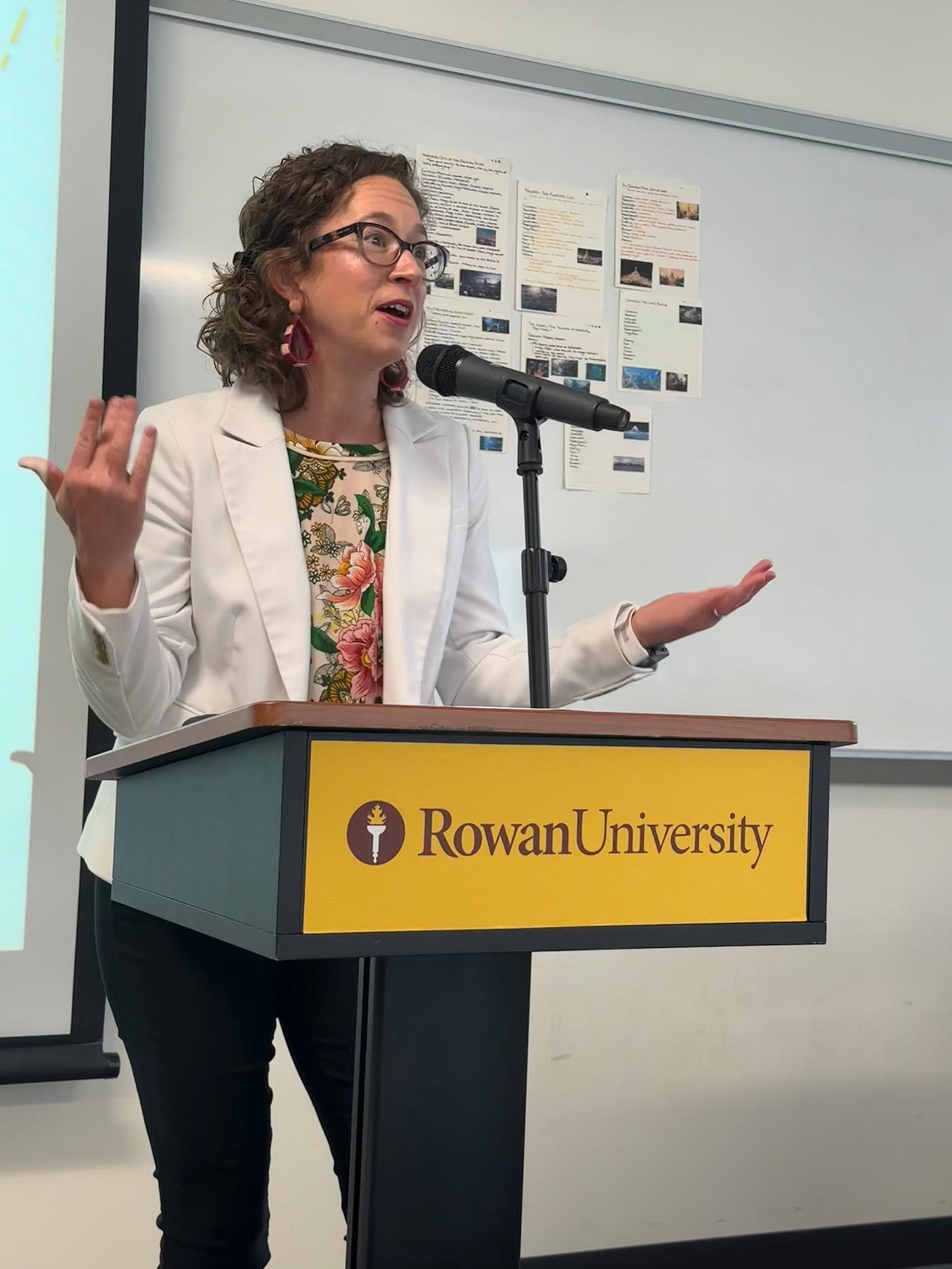 Heather stands at a podium that reads, “Rowan University,” staring past the camera to her class, her hands raised with palms up as she speaks. She wears dark pants, a white jacket, flowered shirt, tortoise shell glasses. On the board behind her, are some posters