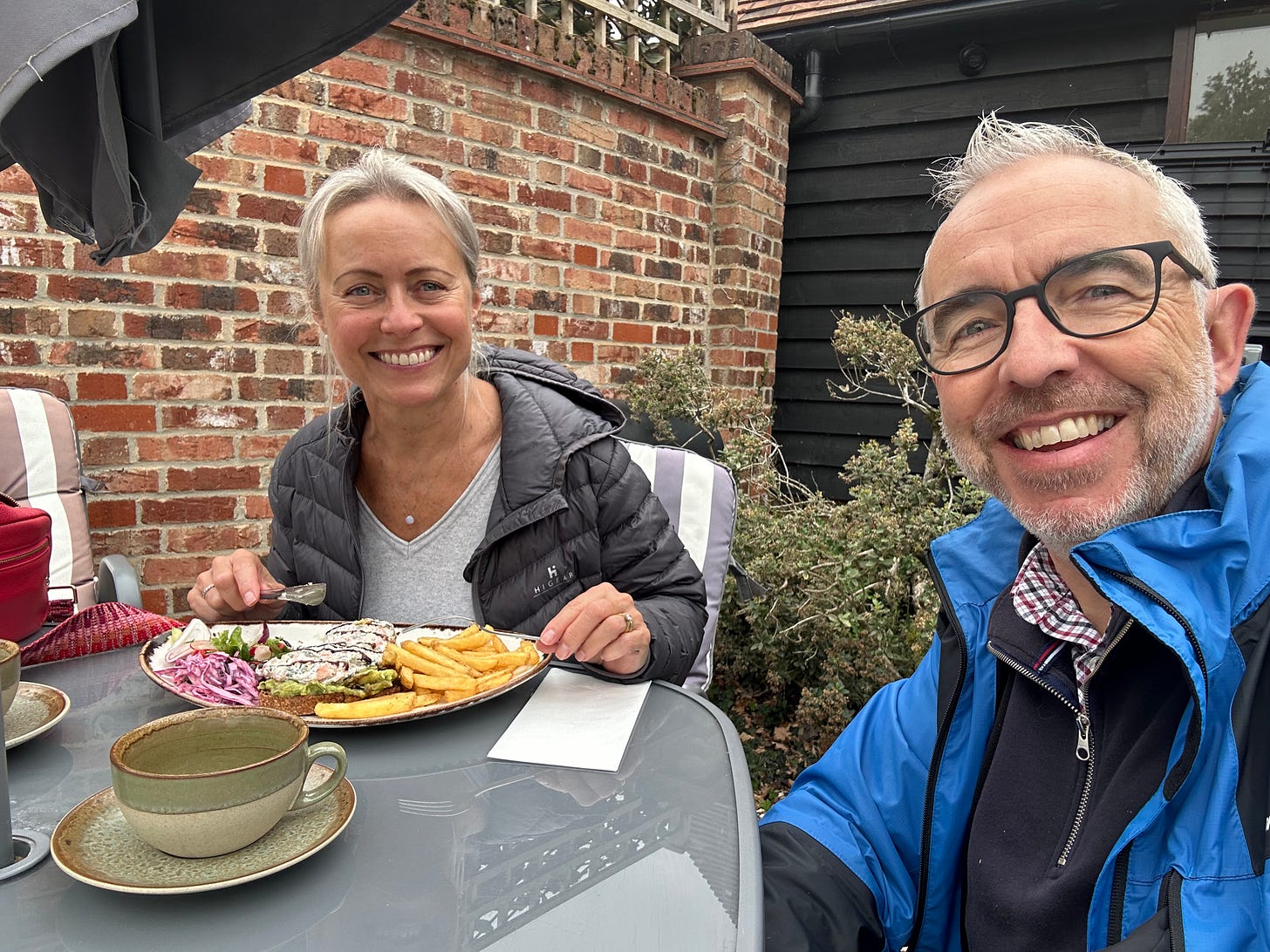 Andy and Julia enjoying lunch outdoors at a café table on a cool day, with a brick wall and dark wooden building in the background.