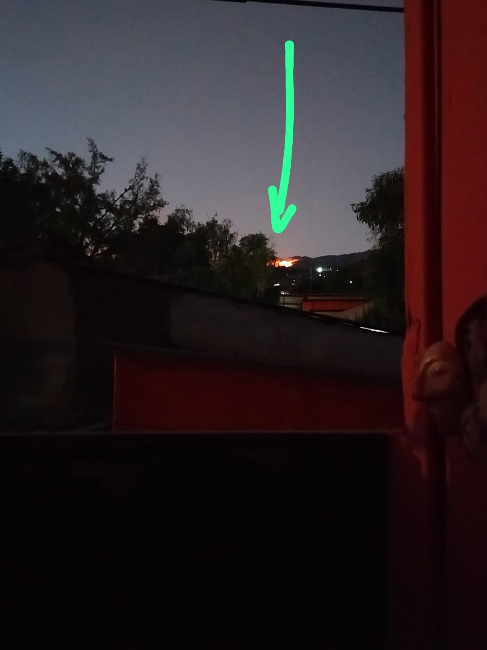 A photo, taken at night, showing a fire has broken out on a hill in the distance. there is an arrow drawn on the photo pointing to the location of the fire.
