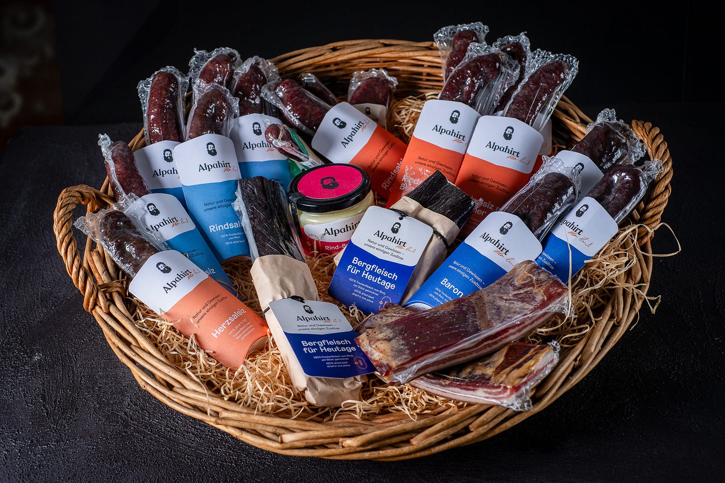 Ein geflochtener Präsentkorb ist prall gefüllt mit verschiedenen Trockenfleisch- und Wurstprodukten der Marke «Alpahirt». In der Mitte stehen vakuumierte Stücke Bergfleisch und ein Glas mit rotem Deckel, aussen herum zahlreiche verpackte Würste und Fleischstücke mit farbigen Etiketten in Blau, Weiss und Orange. Als Unterlage dient Holzwolle, der Hintergrund ist dunkel gehalten.