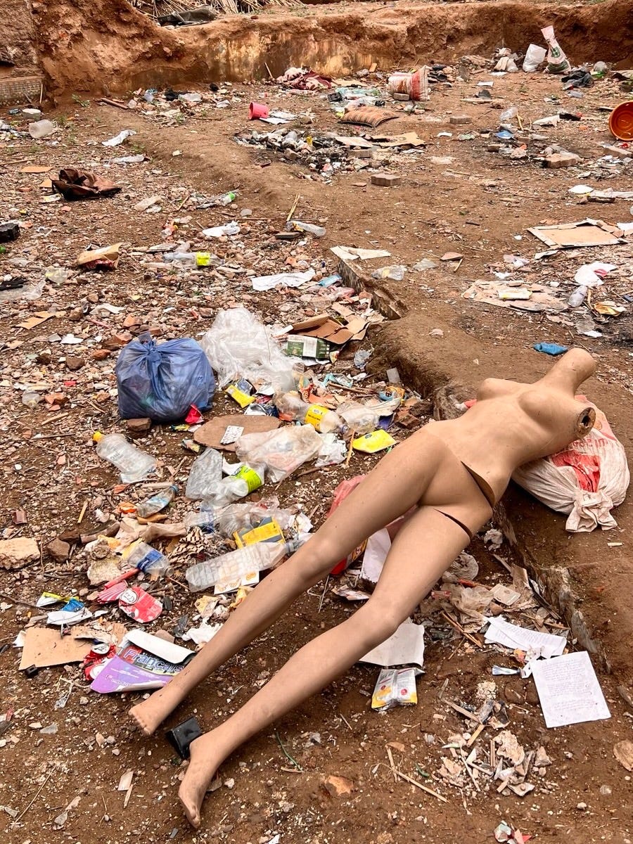 A broken mannequin torso and legs lie discarded among trash and rubble on a dusty ground, surrounded by plastic bags, bottles, and debris. A broken mannequin torso and legs lie discarded among trash and rubble on a dusty ground, surrounded by plastic bags, bottles, and debris.