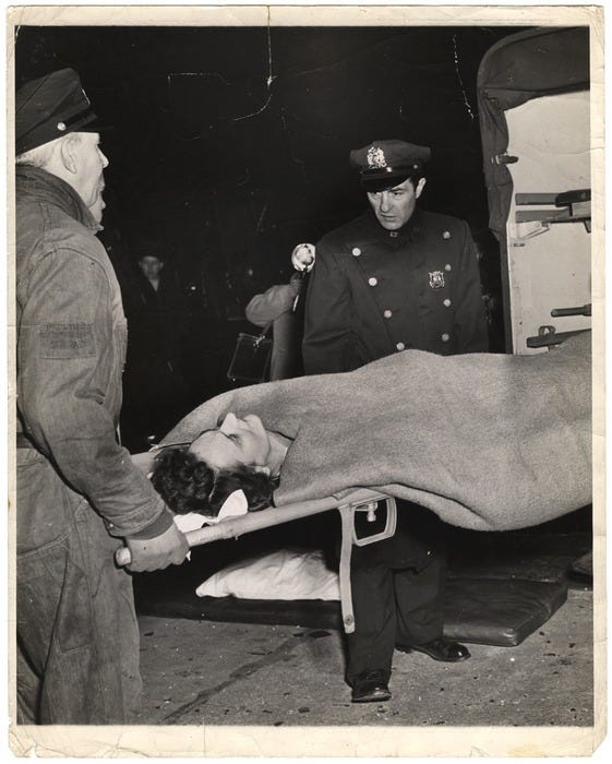 [Police officer looking at woman on stretcher]