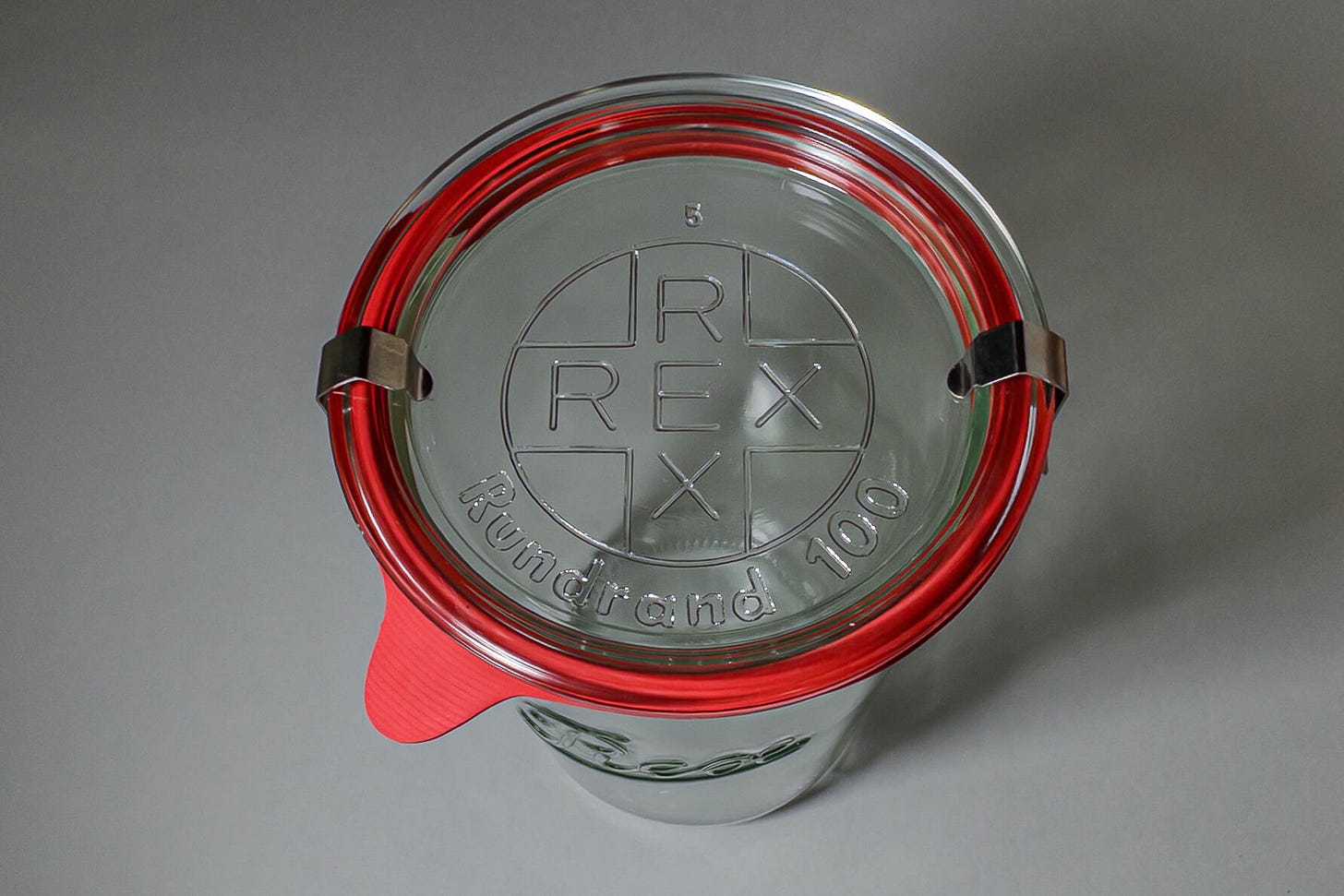 Draufsicht auf ein geschlossenes REX-Rundrandglas mit Glasdeckel, orangem Gummiring und zwei Metallklammern; auf dem Deckel ist das eingeprägte REX-Logo in Kreisform sowie die Aufschrift „Rundrand 100“ zu sehen. Draufsicht auf ein geschlossenes REX-Rundrandglas mit Glasdeckel, orangem Gummiring und zwei Metallklammern; auf dem Deckel ist das eingeprägte REX-Logo in Kreisform sowie die Aufschrift „Rundrand 100“ zu sehen.