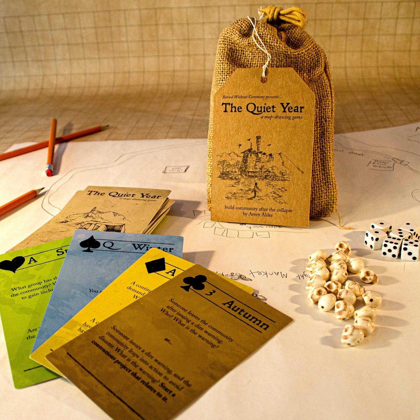 A bag and colored cards with traditional playing card suits and numbers, as well as prompts and game-mechanics written on them. All on top of a hand-drawn paper map.