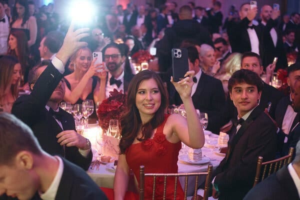 A woman in a red dress holds up her phone as a speech is delivered. She is surrounded by people at tables, many of them also recording the moment with their phones.