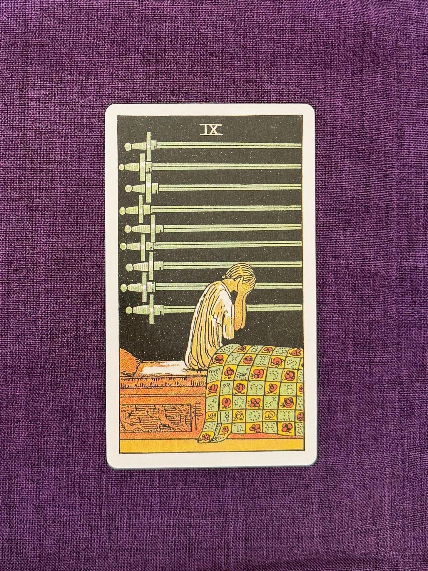 The Nine of Swords shows a woman sitting up in her bed with her head in her hands. Nine swords hang on the dark wall behind her. The base of the woman’s bed features a carving of one person defeating another.