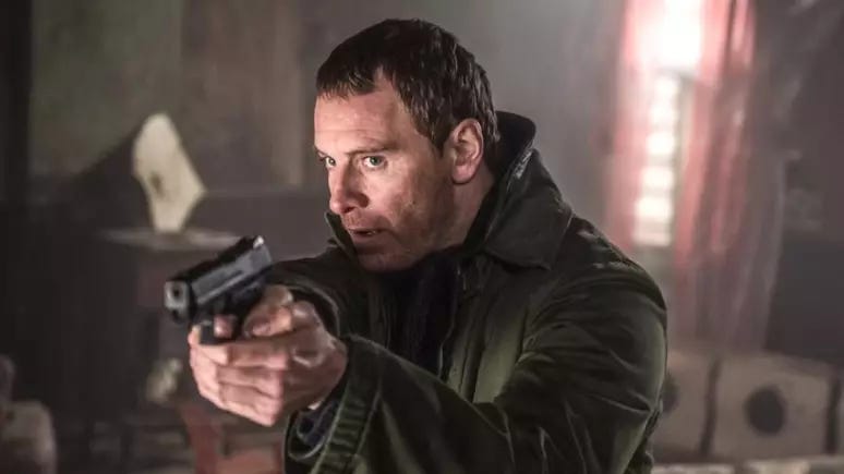 Foto de Michael Fassbender em cena no filme O Assassino. Ele aponta uma arma a uma pessoa fora do quadro da imagem e tem um olhar ameaçador. Foto de Michael Fassbender em cena no filme O Assassino. Ele aponta uma arma a uma pessoa fora do quadro da imagem e tem um olhar ameaçador.