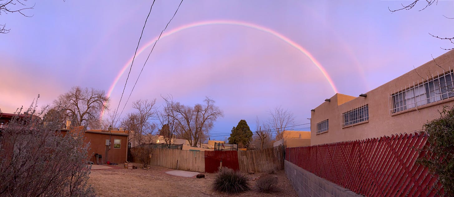 A large rainbow in a pale blue morning sky over adobe style homes