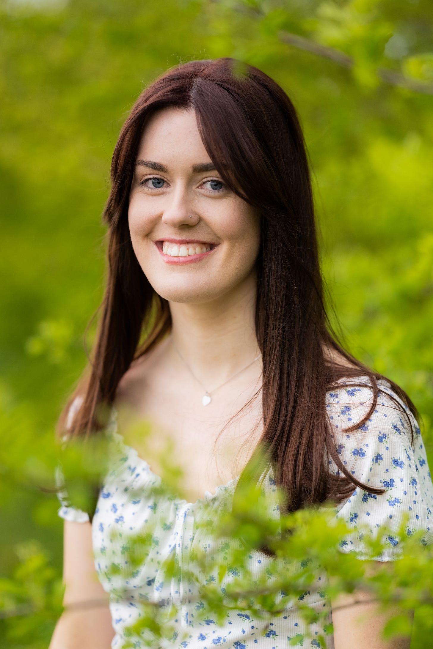 Emily, a young white woman with reddy brown hair and bangs is smiling at the camera. She is wearing a white top with small blue flowers on it. She is surrounded by greenery.