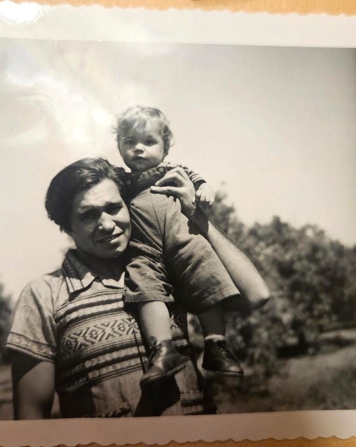 Black and white photo of Winona's dad holding baby Winona in his left hand as he looks at the camera. He is wearing a patterned short sleeve collared shirt. She is wearing a jumpsuit. He has black hair, parted on the side. She has blond hair.