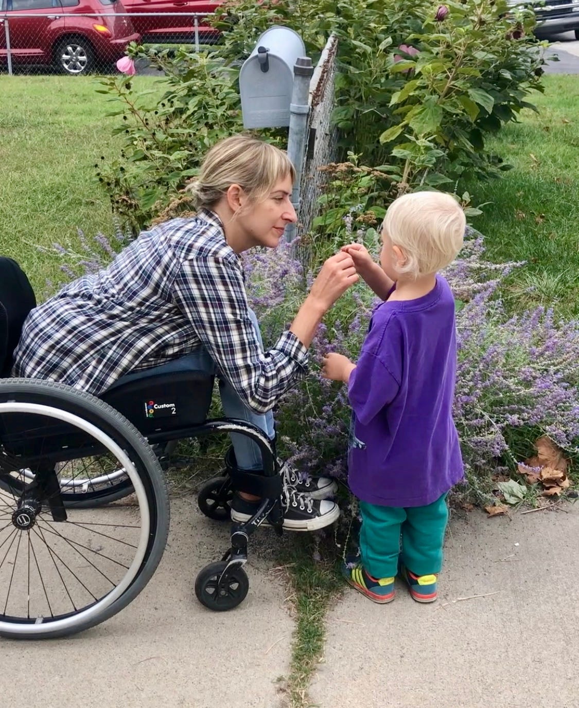 Rebekah and her son huddled up next to a lavender bush growing out of the side of a chain link fence. Rebekah sits in her wheelchair facing her two-year-old son who’s holding out a stem for her. 