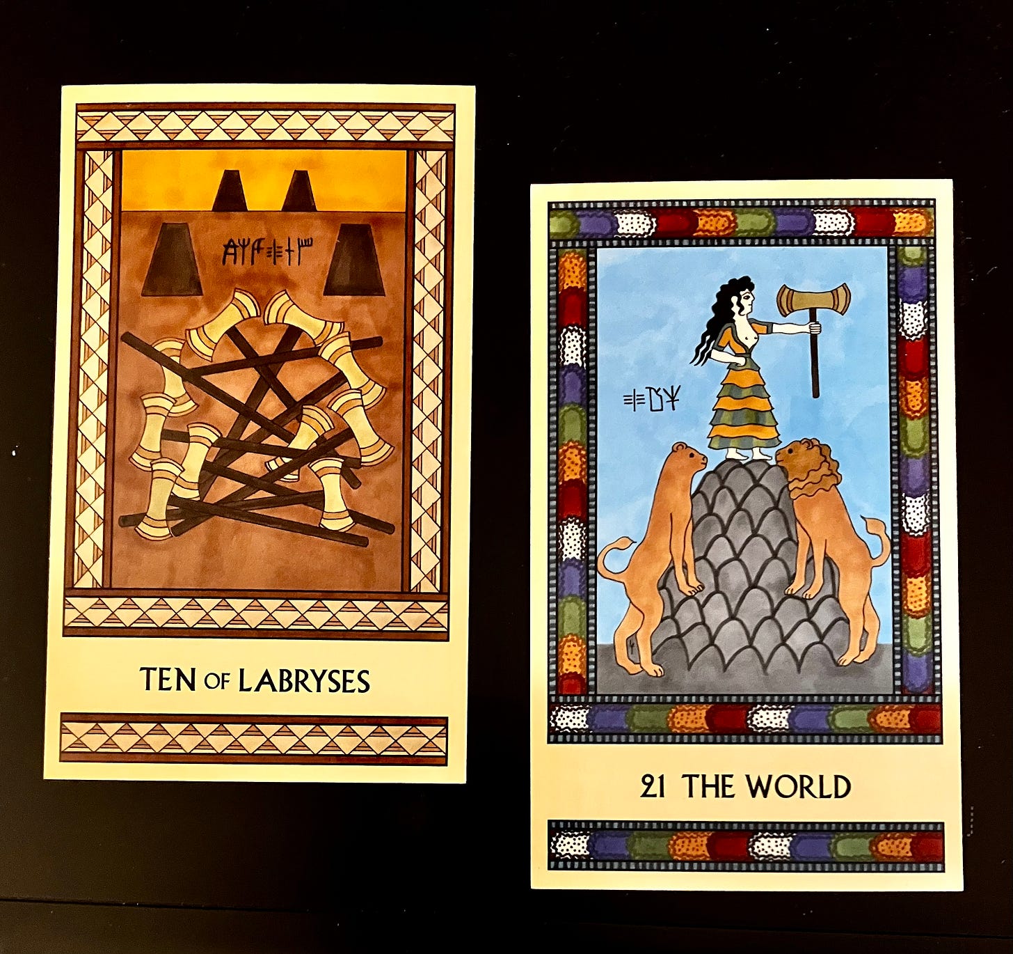 Two Minoan Tarot cards side by side on a black surface. The Ten of Labryses is painted in shades of gold, cream, and tan. It shows ten labryses tangled in a heap on the floor while four empty labrys stands sit in the background. The World has a bright multicolored border. It shows a goddess standing atop a tall mountain, holding a labrys in her outstretched hand. A pair of lions, one female and one male, flank the base of the mountain. 