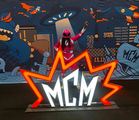 I'm dressed like a pink power ranger posing with my fist up stood behind a neon MCM sign