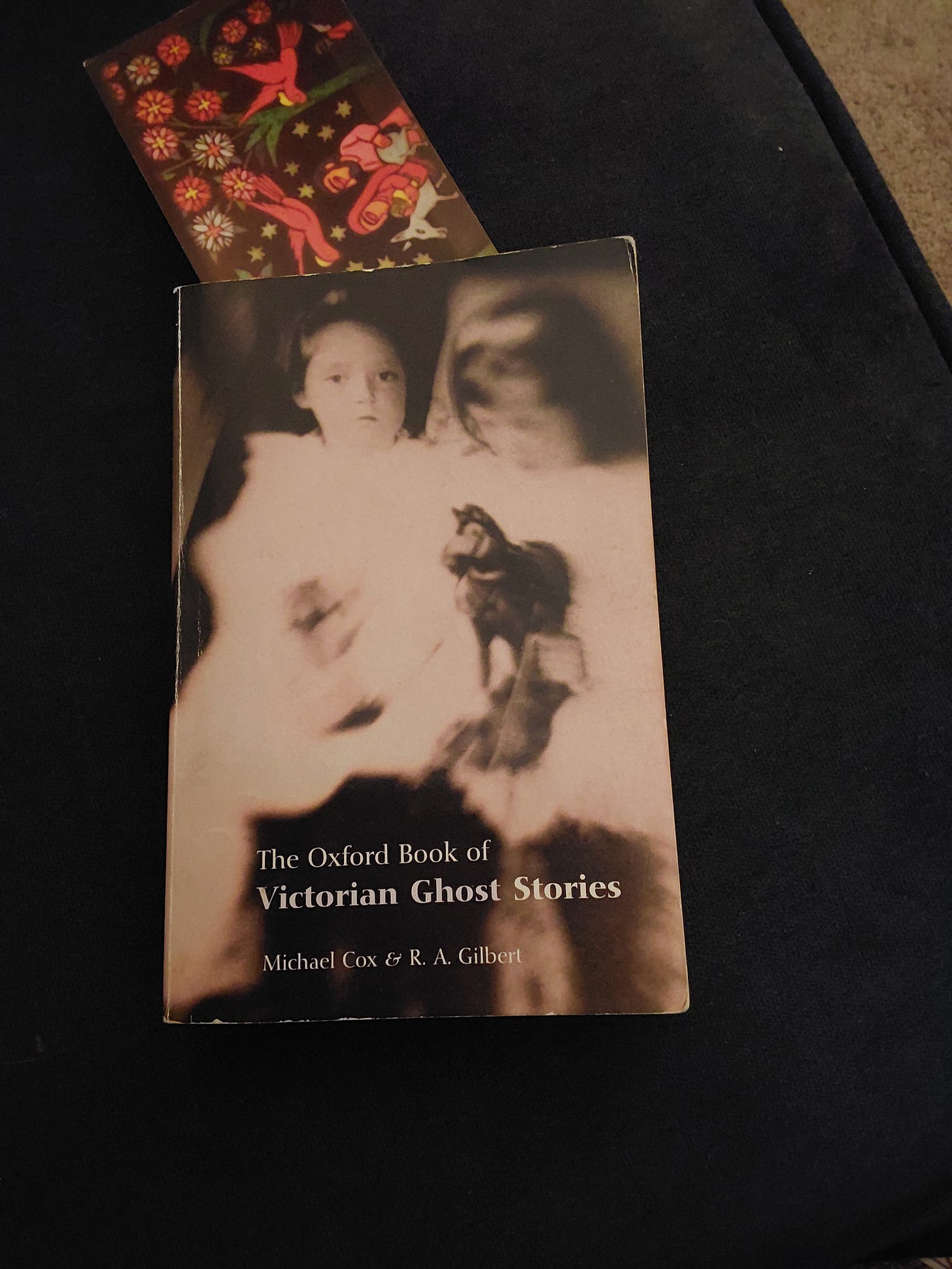 A photo of a paperback book on a black background. The book is titled The Oxford Book of Victorian Ghost Stories, and has a blurred image of two children, one holding a toy horse. Above the book is a bookmark with a design in the medieval style of Mary and Joseph on a donkey.