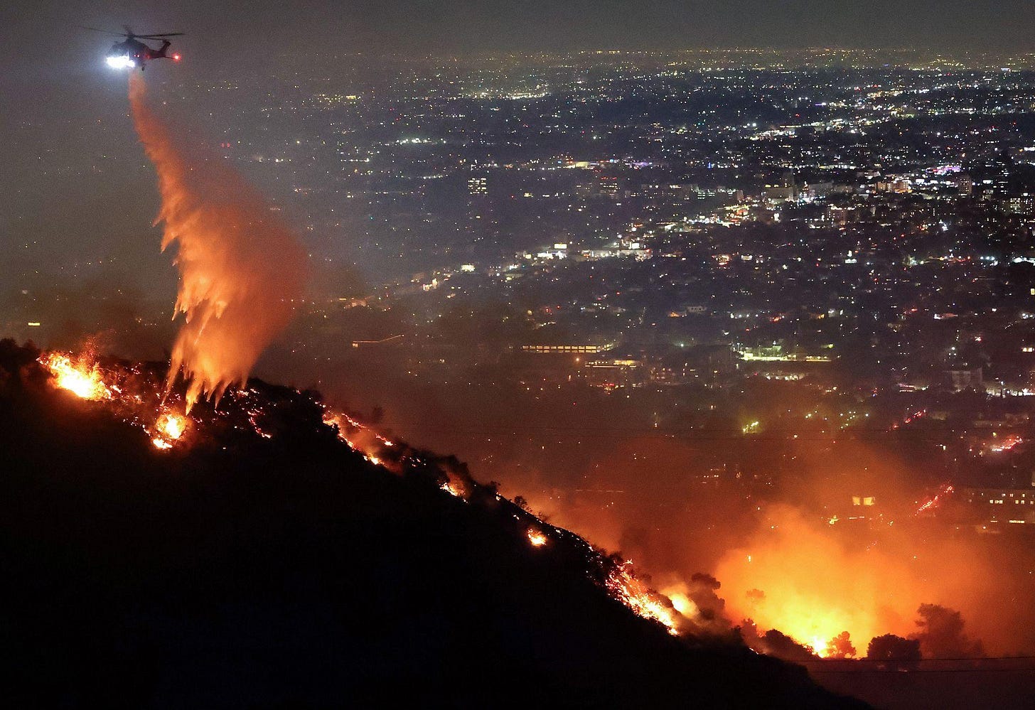 A firefighting helicopter drops water as the Sunset Fire burns in the Hollywood Hills at night time on Wednesday, with a view of the LA skyline in the background. A firefighting helicopter drops water as the Sunset Fire burns in the Hollywood Hills at night time on Wednesday, with a view of the LA skyline in the background.