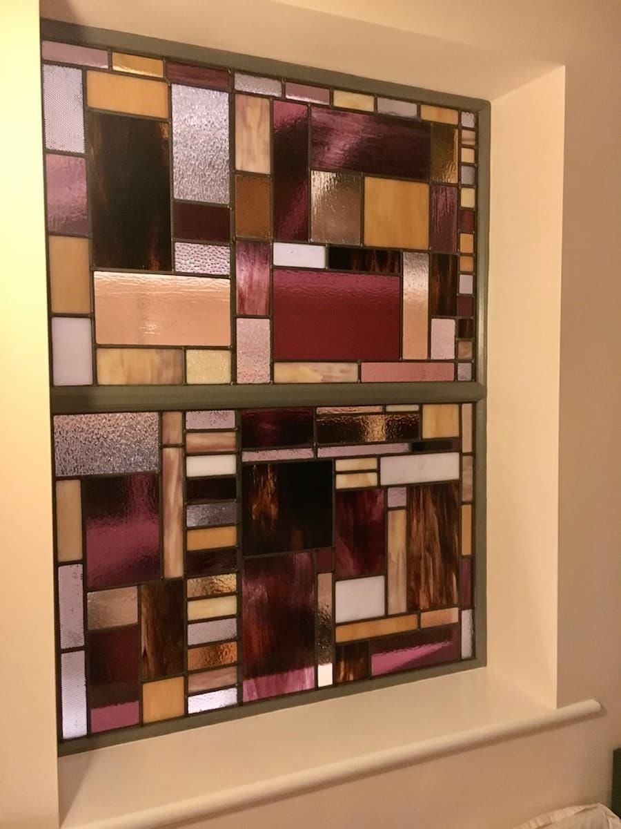 Stained glass window, the design is made up of small, rectangular and square panels in various colours and textures. The colours include shades of pink, red, gold, brown, and clear glass. 