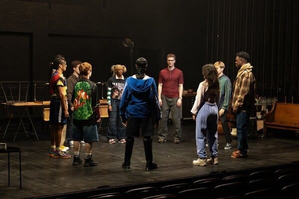 Nine people stand in a circle onstage in this production image. Nine people stand in a circle onstage in this production image.