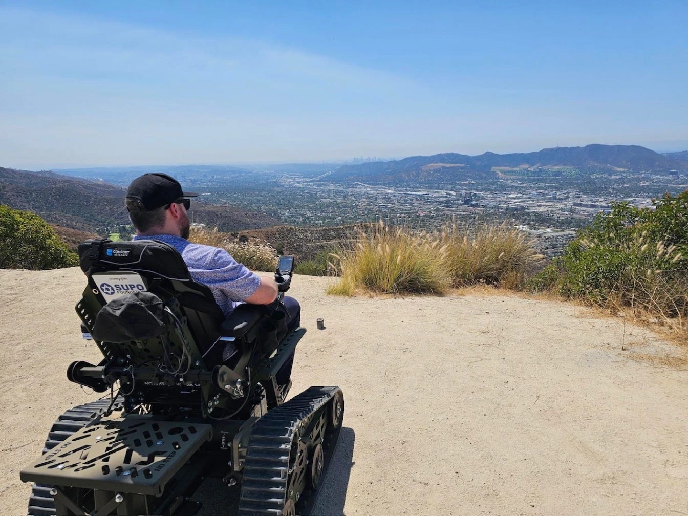 A man in a heavy-duty off-road wheelchair is looking out over a huge vista of land during a sunny day on a dirt path.