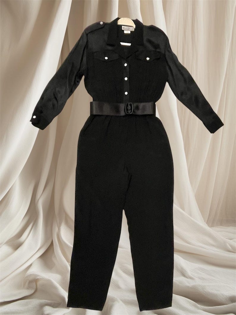 May include: A black satin jumpsuit with a collared shirt, long sleeves, and a black belt. The jumpsuit has two flap pockets on the chest and a button closure in the front.