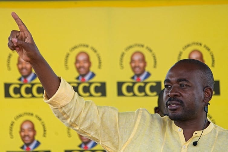 A man points ahead with his right index finger in front of banners bearing the acronym 'CCC'. A man points ahead with his right index finger in front of banners bearing the acronym 'CCC'.