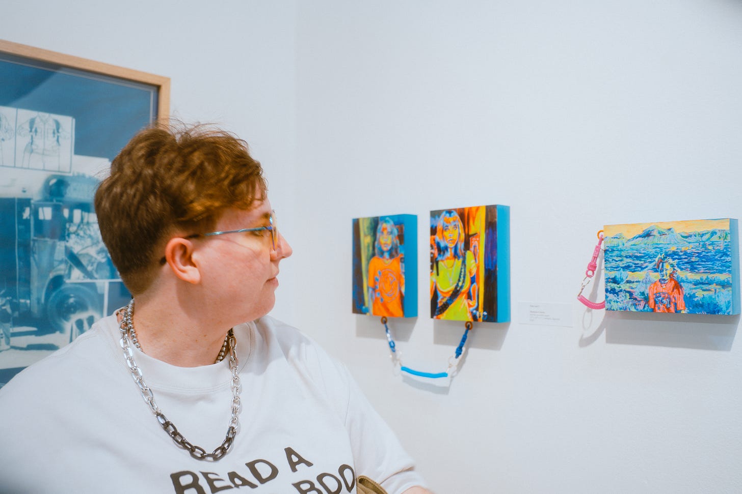 Non-binary person in foreground looking to the right at their artwork hanging on the wall, displayed in a gallery.