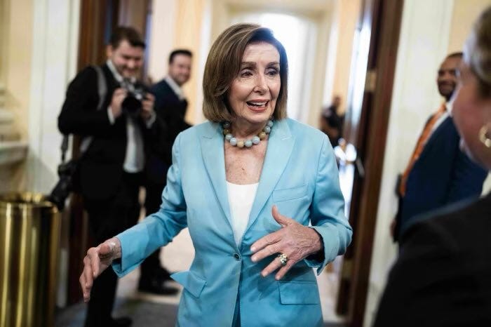 Nancy Pelosi stands in a congressional hallway wearing a light blue suit jacket and white top with a pearl necklace, gesturing with her hands while smiling slightly. Surrounding her are several men in dark suits, some holding cameras and notebooks, appearing to be press members. The setting includes wooden doors and a trash can in the background.