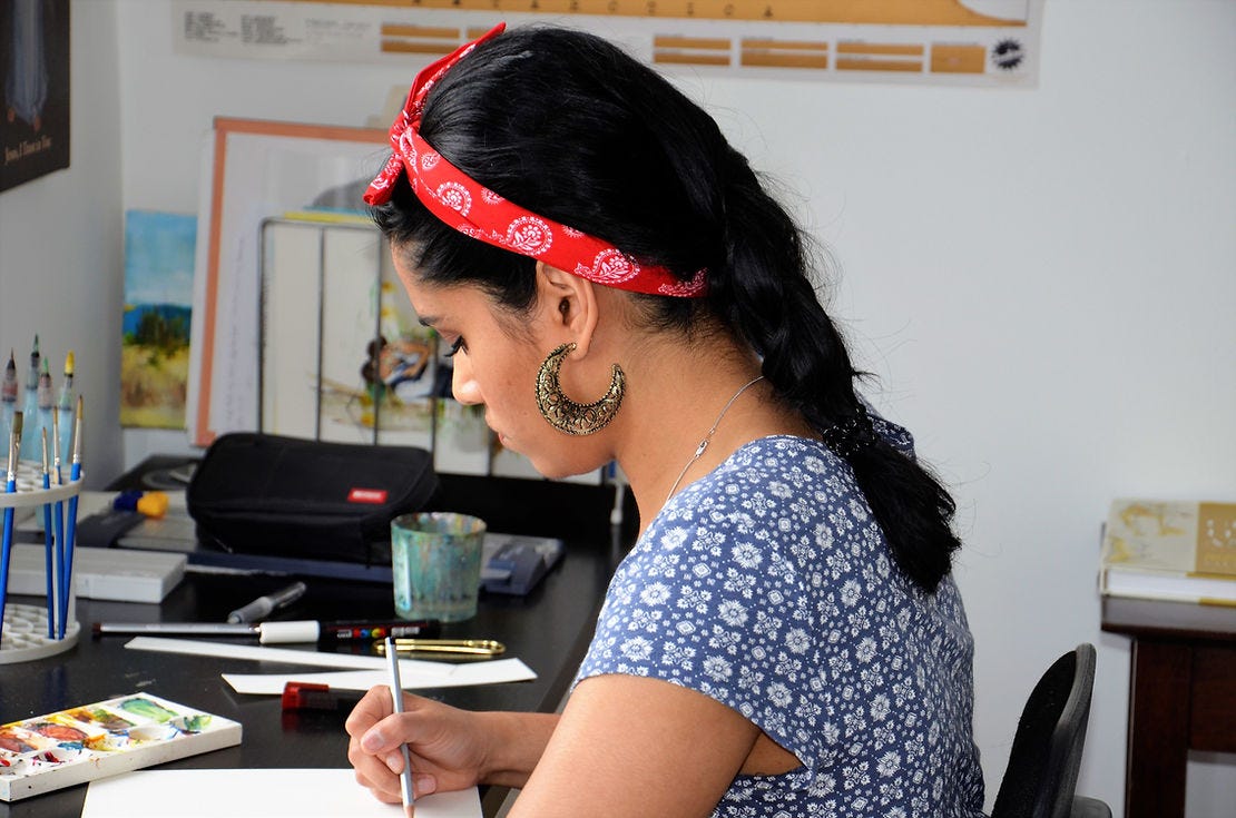 There is a side profile of guest Trisha de Souza. She is in the midst of drawing. In the foreground is her desk that has various art supplies strewn about.