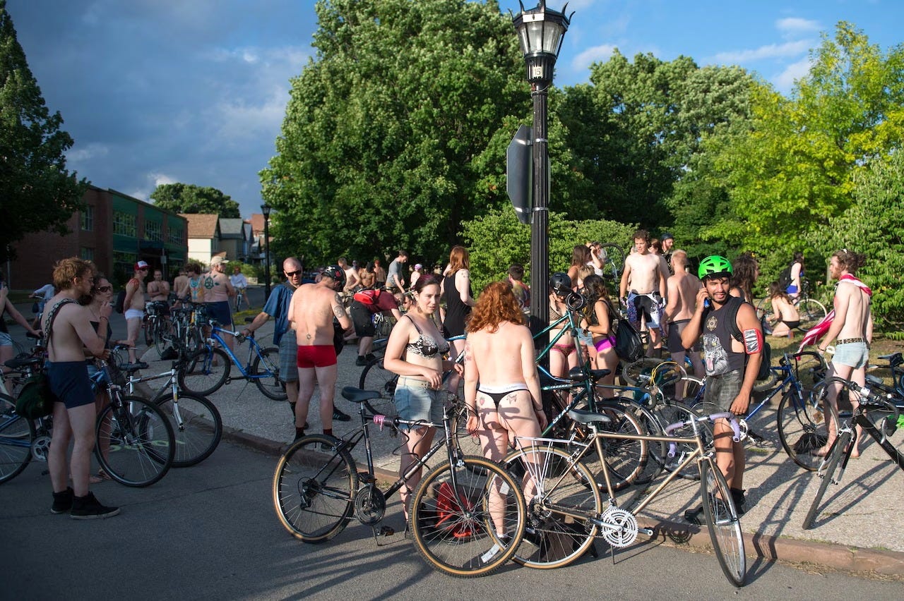 Cyclists in various states of undress gather with their bikes at a tree-lined street corner beneath a streetlamp, talking and waiting for a ride to begin. Cyclists in various states of undress gather with their bikes at a tree-lined street corner beneath a streetlamp, talking and waiting for a ride to begin.