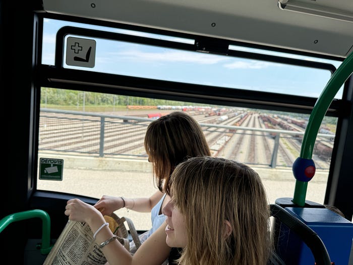 Two teen girls riding the bus, with old trains on tracks outside the window behind them. Two teen girls riding the bus, with old trains on tracks outside the window behind them.