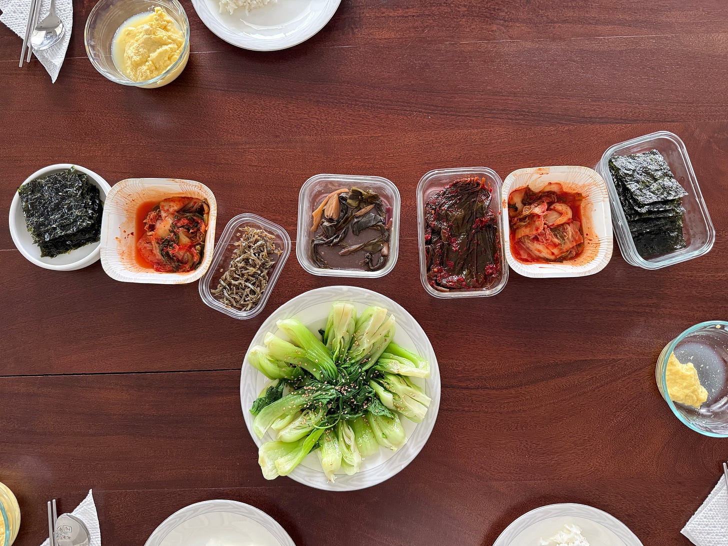 Overhead view of a Korean dinner table with banchan — kimchi, seaweed, steamed greens, marinated leaves, dried anchovies, and rice — laid out in small dishes for everyone to build their own plate.