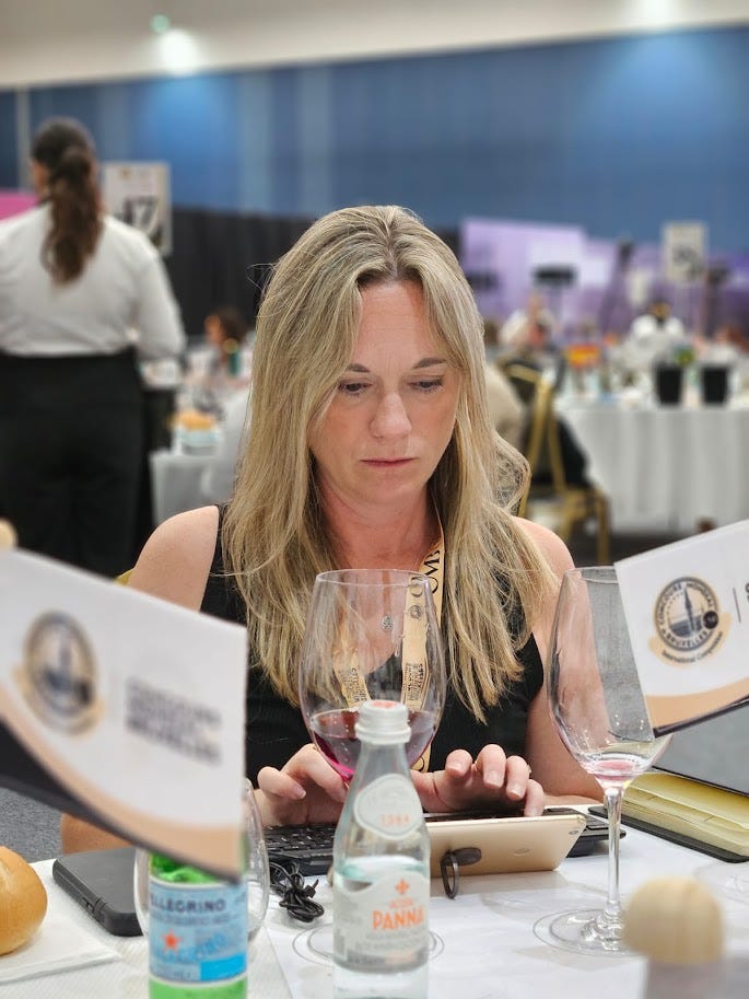 Kristy focused on entering tasting notes at a Concours Mondial de Bruxelles judging table, wine glasses and a keyboard set up in front of her