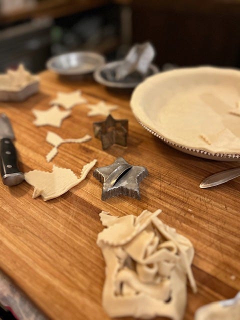 A white cake with a red ribbon ted around it and sprig of pine as the centerpiece; an empty pie shell with cookie cutters and scraps of pie crust on a wooden cutting board.