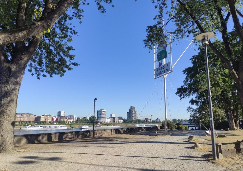 Riverside view, with a sign high up saying "Rheinpark Tanzbrunnen" to welcome everyone. A large river cruiser is moored on the far side of the river, with Cologne's skyline behind it.