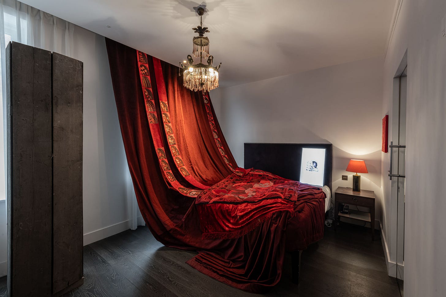 Dim hotel room installation with a dramatic red fabric draped from ceiling to floor, transforming the space into a sculptural environment.