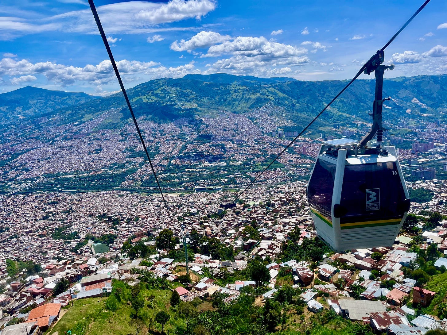 A Metrocable gondola climbs above Medellín's hillside neighborhoods, the city filling the Aburrá Valley with green mountains beyond. A Metrocable gondola climbs above Medellín's hillside neighborhoods, the city filling the Aburrá Valley with green mountains beyond.