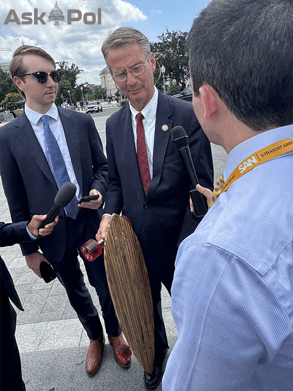 A man in a suit talks to reporters outside while holding a longboard. Photo Matt Laslo for © www.askapoluaps.com