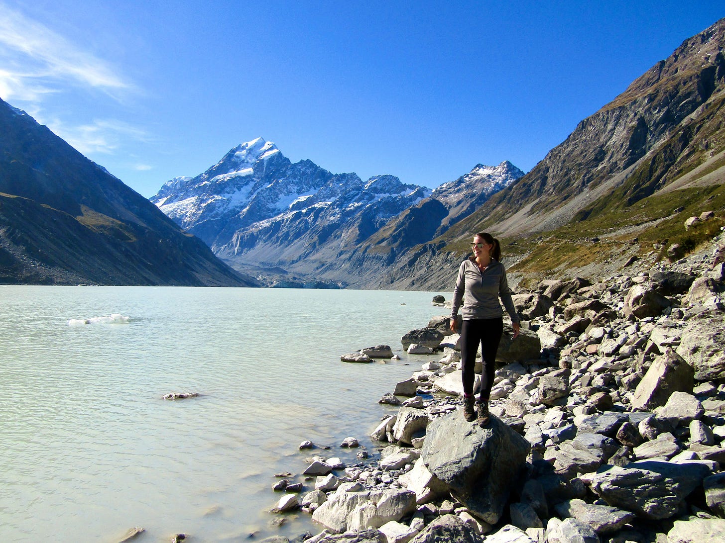 A woman stands on rocky shores beside a milky glacial lake, with snow-capped mountains rising dramatically behind her under a deep blue sky. A woman stands on rocky shores beside a milky glacial lake, with snow-capped mountains rising dramatically behind her under a deep blue sky.