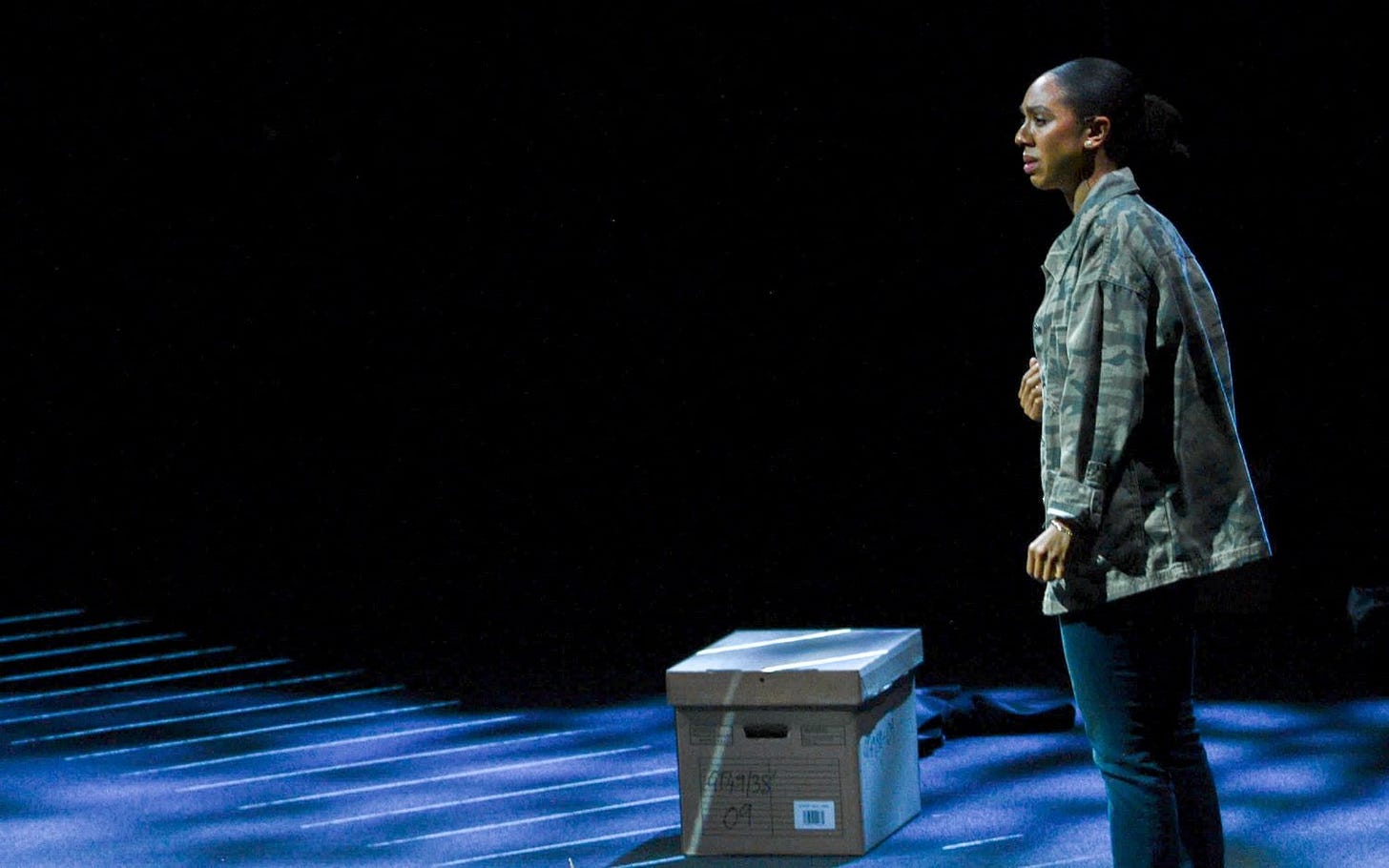 A woman in a camouflage jacket stands, with a brown cardbox box on the floor.