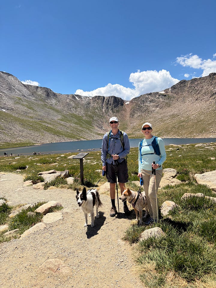 a man, woman and two dogs standing in front of a lake surrounded by mountains and a man and two dogs on a rocky trail