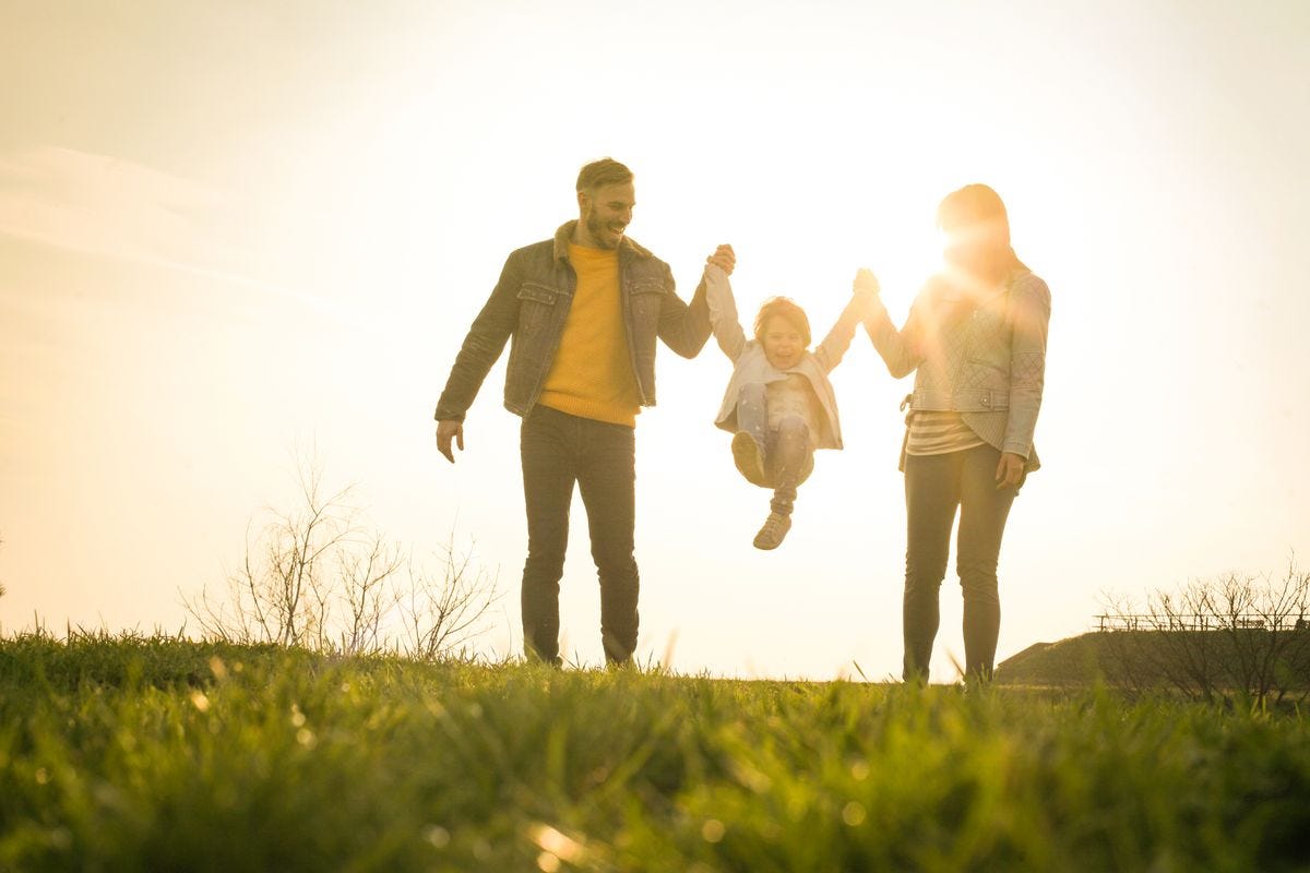 Happy parents walking in nature swinging their daughter between them, backlit by sun.