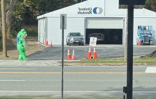 A person wearing a frog costume walks on a sidewalk. A person wearing a frog costume walks on a sidewalk.