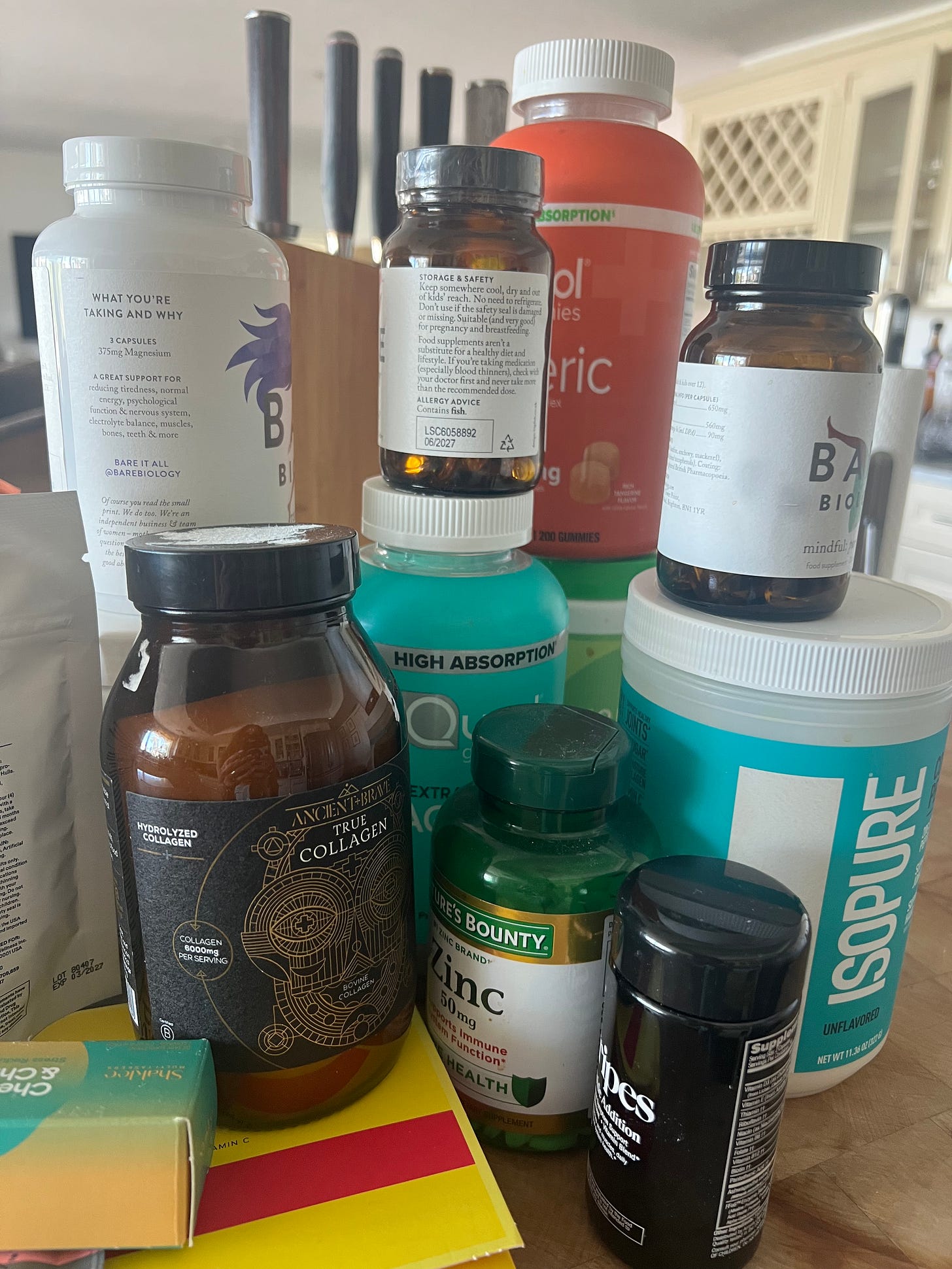 A variety of beauty and health supplements, including collagen, magnesium, and zinc, piled on a kitchen counter. The image depicts "supplement overwhelm," featuring brands like Bare Biology, Isopure, and Ancient + Brave.