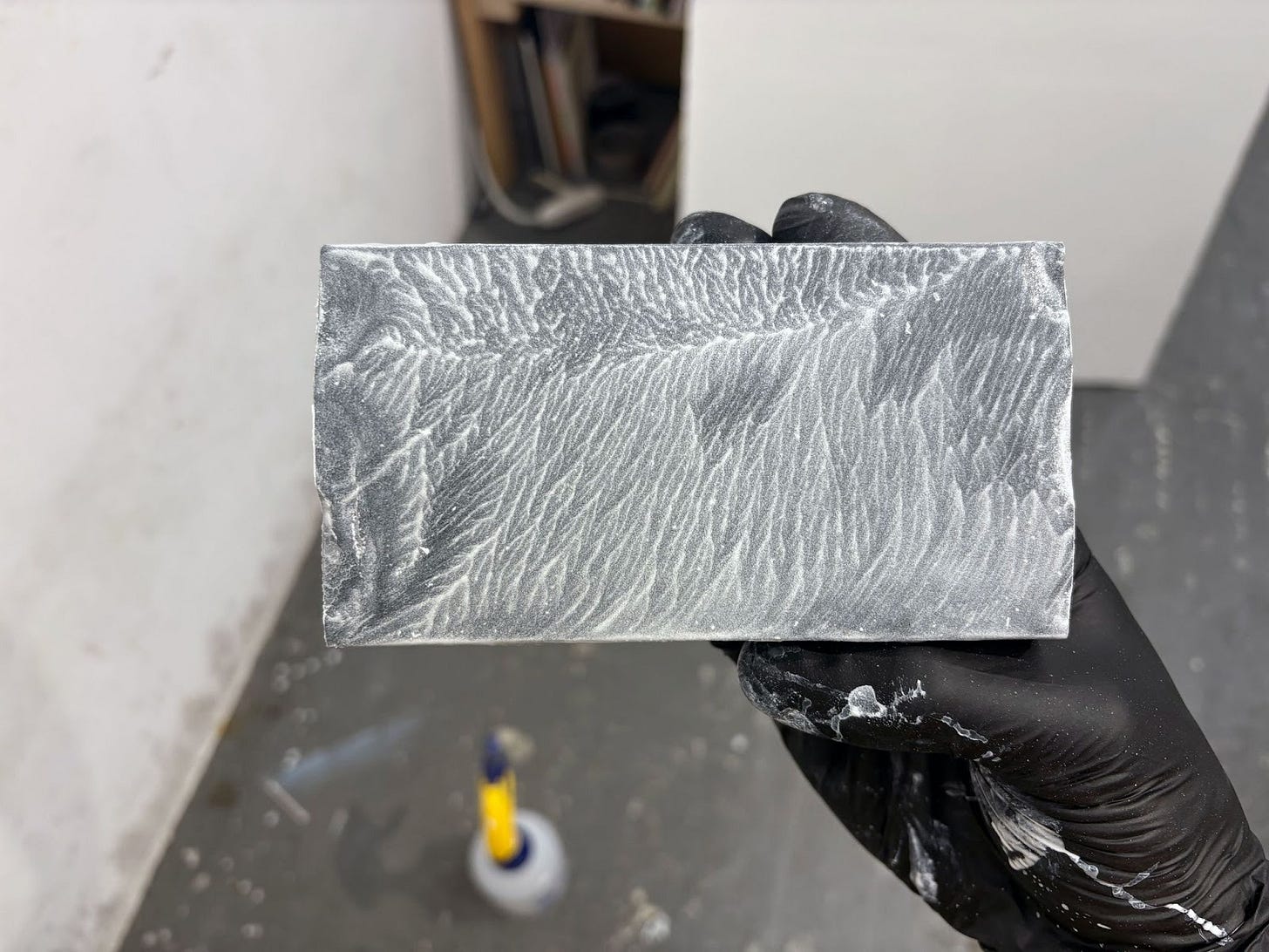 A hand in a black nitrile glove holds a sanding block that is covered in a wet, white slurry.