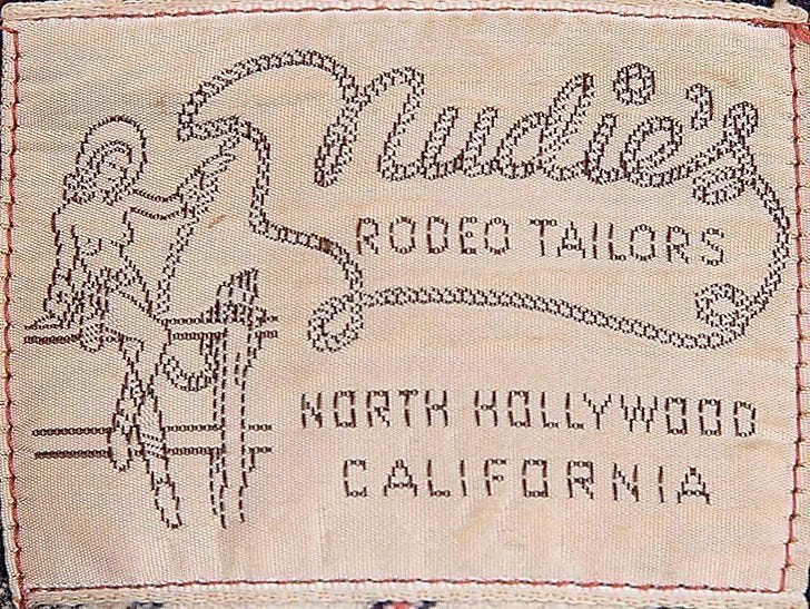 The original tag for Nudie's Roder Tailors. The naked cowgirl motif arose when Nudie's wife Bobbie appeared in the doorway wearing only a hat, boots, and a holster, asking him when he was going to make the rest of the outfit.