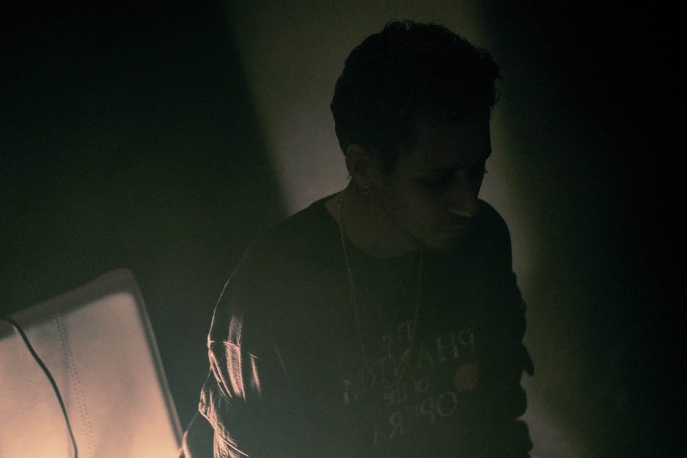 cøzybøy is standing and posing facing the bottom right corner. He is shrouded in darkness with a soft light source on the bottom left.
