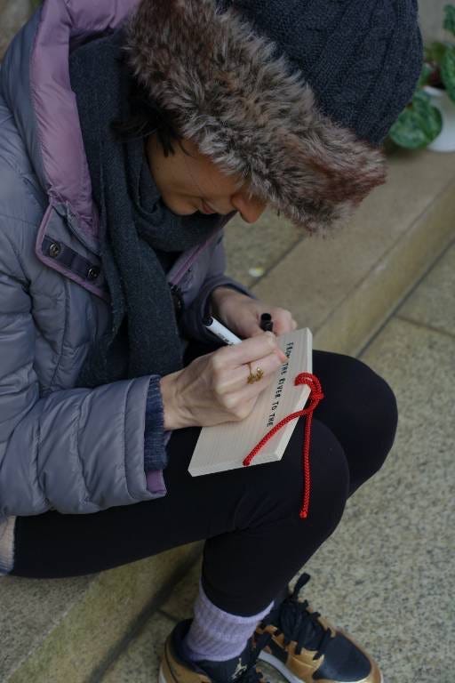 a woman wearing a jacket writes something on a small wooden board with a red string through the top