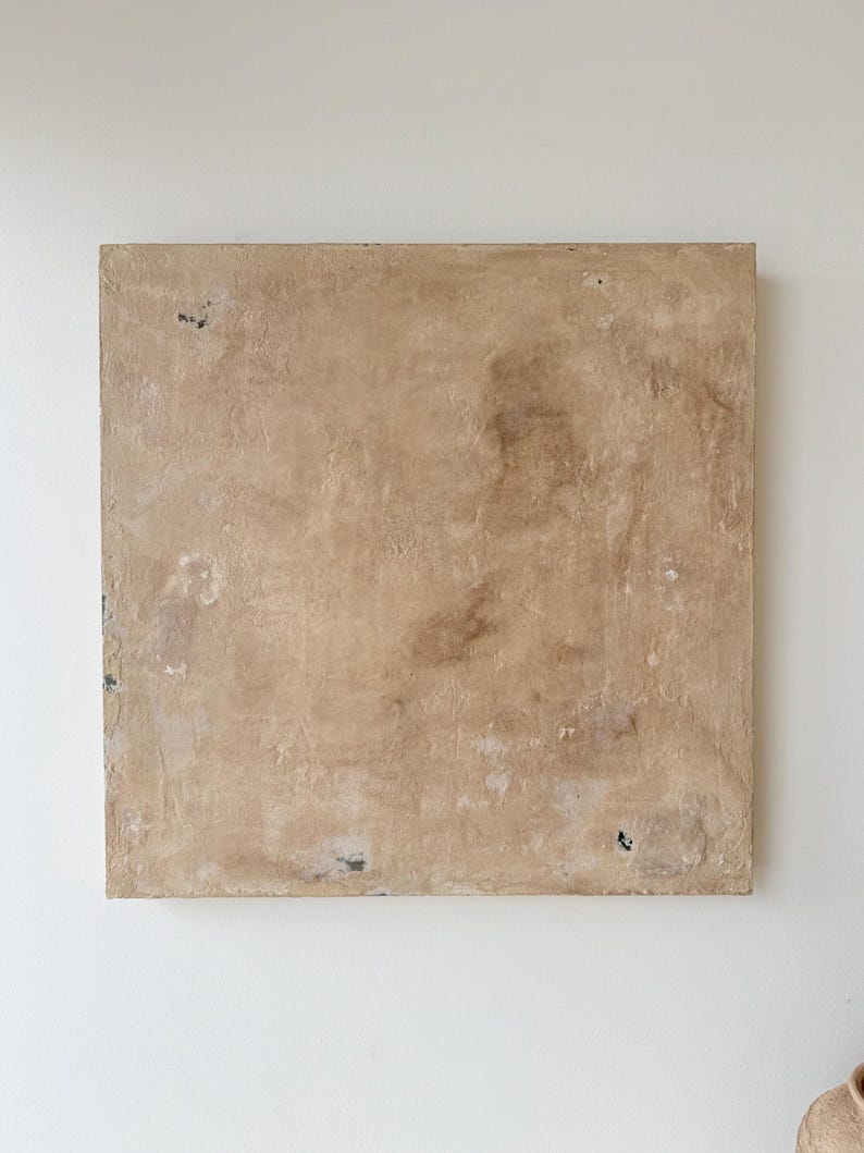 May include: A square, textured, beige-colored wall art piece. The surface has a mottled appearance with darker brown and white patches. The edges show slight wear and tear, adding to its rustic aesthetic. The artwork is mounted on a white wall.