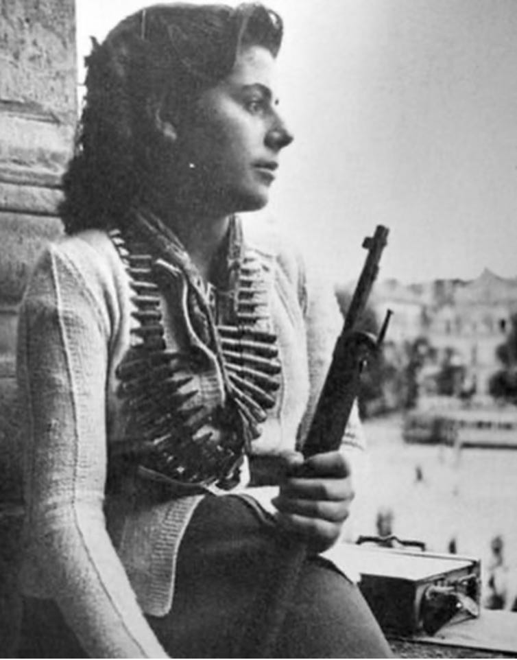 A faded black and white photo of a white woman holding a rifle with bullets hanging around her neck. Beside her is a radio. 