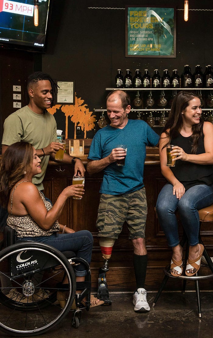A group of people at a bar. One is in a wheelchair, one has a prosthetic leg, and others are sitting down.
